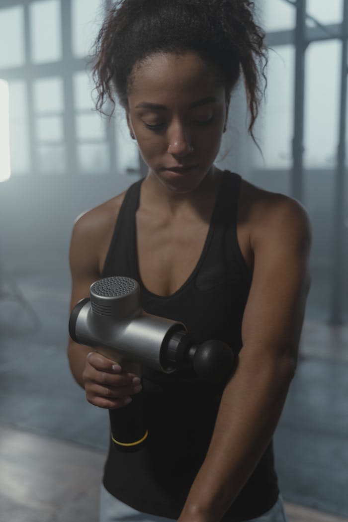 A focused woman using a massage gun on her arm in a gym environment.